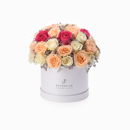 White box filled with vibrant pink, peach, and cream roses accented with delicate purple filler.