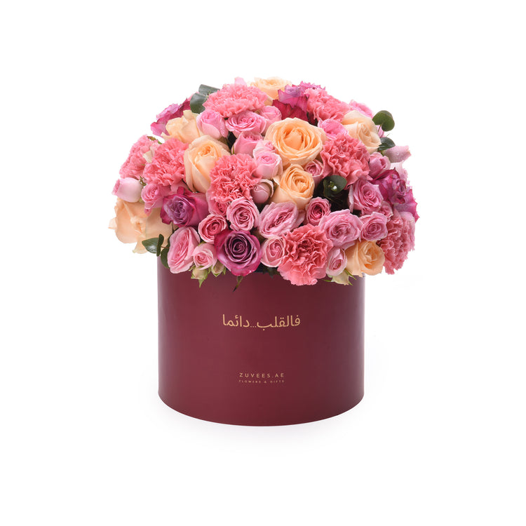 Vibrant pink, peach, and magenta roses and carnations overflowing from a deep burgundy hat box with gold Arabic script.