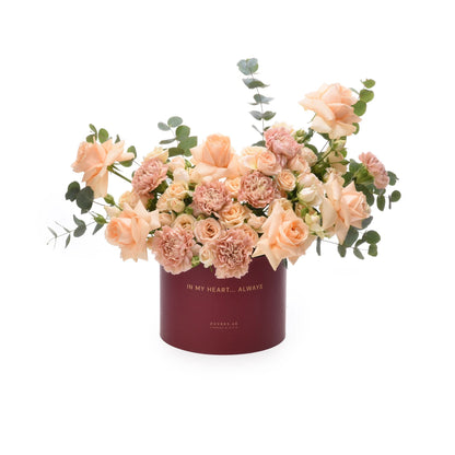 Soft peach roses and carnations in deep burgundy box with gold text.